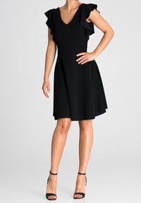 Vestido negro con mangas cortas y volantes, y escote en V. Forma acampanada en A, hasta la rodilla, combinado con tacones negros con tiras. Textura suave.