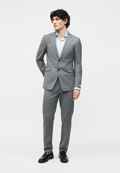 Νέος άνδρας που στέκεται, φοράει γκρίζο κοστούμι slim-fit με αντίστοιχο παντελόνι, γαλάζιο πουκάμισο και μαύρα δερμάτινα loafers, σε λευκό φόντο.
