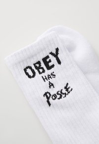 Calcetines blancos acanalados con texto negro que dice "OBEY TIENE UN Posse", con una tela texturizada y forma de calcetín estándar.