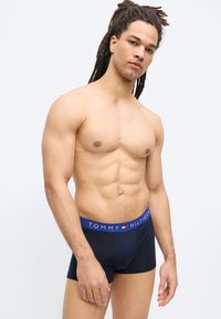 Μυώδης άνδρας με dreadlocks που φοράει μπλε σκούρο ανδρικά εσώρουχα Tommy Hilfiger, στέκεται μπροστά σε ένα απλό φωτεινό φόντο.