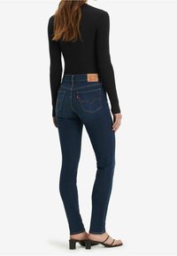 Vaqueros pitillo en denim azul oscuro con cintura alta. Presentan bolsillos traseros clásicos y una etiqueta de cuero con el logo en la cintura. Textura suave.