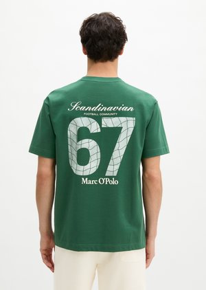Muž v zelenej tričku s textom „Scandinavian Football Community 67 Marc O'Polo“ a sieťovým vzorom na chrbte stojí na bielom pozadí.