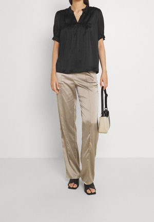Blusa di raso nera con maniche corte a palloncino e scollatura a V, abbinata a pantaloni di raso beige chiaro. Tacchi aperti neri e una piccola borsa.