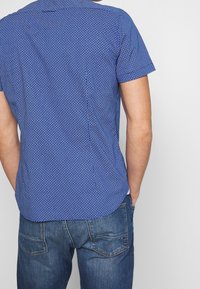 Camicia a maniche corte blu con motivo geometrico, caratterizzata da un orlo arrotondato. Abbinata a jeans in denim blu con tasche posteriori.