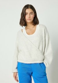 Weißer, flauschiger Pullover mit Wickeldesign, langärmlig und mit Ausschnitten. Kombiniert mit blauen, strukturierten Jogginghosen.