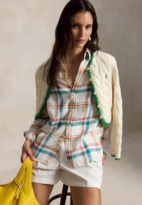 Kvinne iført en kremfarget kabelstrikket cardigan over en flerfarget rutete skjorte med hvite shorts, holder en gul håndveske, sittende på en krakk.