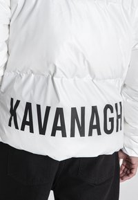 Hvid polstret jakke med en blank finish, udstyret med stor sort skrift "KAVANAGH" på ryggen, og kontrasterende sorte bukser.