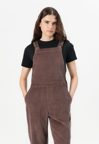Salopette in corduroy marrone con costine verticali, dotata di bretelle regolabili, tasche laterali e una vestibilità comoda. Indossata sopra una maglietta nera.