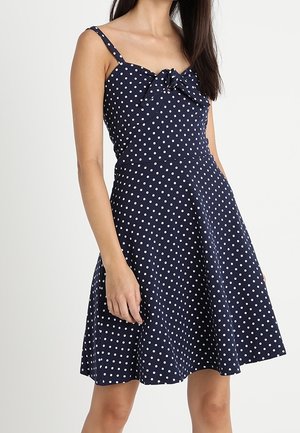 Vrouw draagt een marineblauwe mouwloze jurk met witte polkadots en een strikdetail aan de voorkant, staand tegen een effen lichte achtergrond.