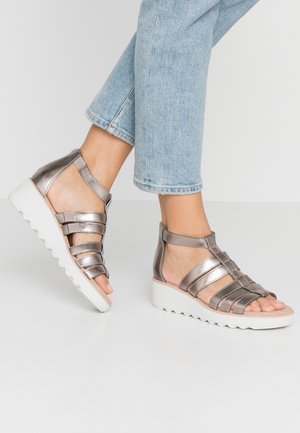 Fødder iført sølvfarvede kilehælssandaler med hvide såler kombineret med lyseblå cropped jeans mod en ensfarvet baggrund.