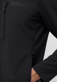 Schwarze Softshell-Jacke mit einer Reißverschlusstasche, die ein Logo trägt, eine glatte Textur hat und tailliert geschnitten ist. Reißverschlüsse sind an den Seiten deutlich sichtbar.
