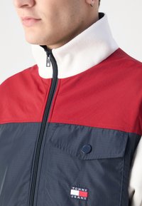 Giacca zip-up rossa e blu navy con colletto bianco, dotata di una tasca frontale, tessuto texturizzato e una patch con logo in basso a sinistra.