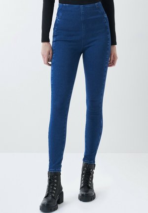 Kobieta w niebieskich rurkach z wysokim stanem i czarnych skórzanych botkach z sznurowaniem, stojąca na tle jednolitego jasnego tła.