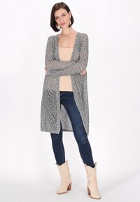 Leichte, offene Strickjacke mit einem schwarz-weißen Muster. Kombiniert mit einem beigen Tanktop, dunklen Jeans und hellen Cowboy-Stiefeln.