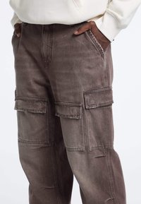 Bruine spijkerbroek cargobroek met twee grote zijzakken, losse pasvorm en lichte vervaging. Heeft een hoge tailleband en gestikte details.