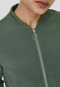 Olive green bomber jacket with a ribbed collar, silver zip closure, and a smooth texture. Simple design with no visible patterns or accents.