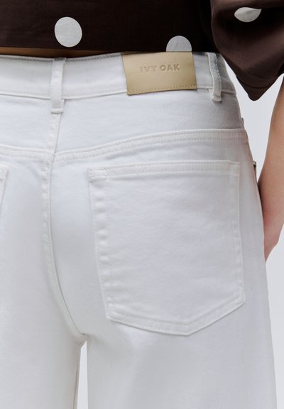 Jean en denim blanc porté par une personne, montrant la poche arrière et l'étiquette beige de la marque "IVY OAK" sur la ceinture, associé à un haut marron à pois.