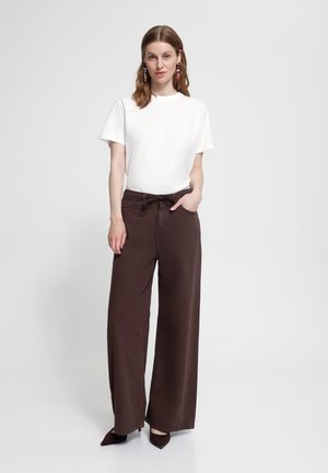 Femme debout devant un fond uni, portant un haut blanc à manches courtes, un pantalon large marron, des talons foncés et des boucles d’oreilles colorées.