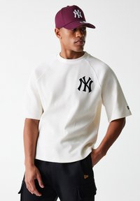 White ribbed short-sleeve shirt featuring a black NY logo on the left chest, paired with a maroon cap with a white NY logo. Black cargo pants.
