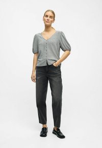 Kvinde står med den ene hånd i lommen, iført en grå kortærmet knapbluse, sorte jeans og sorte loafers foran en ensfarvet baggrund.