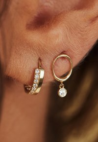 Goldene Creolen mit einer Reihe funkelnder Steine auf einer Seite und einer ovalen Form mit einem danglenden Kristall auf der anderen Seite.