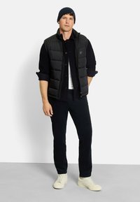 Schwarze Steppweste über einem navy-blauen gerippten Pullover, kombiniert mit navy-blauen Cordhosen und cremefarbenen Sneakers, abgerundet mit einer dunklen Mütze.