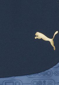 Tejido textil en color navy con un logo dorado de Puma. La sección inferior presenta un diseño azul con patrones que incluyen varios símbolos.