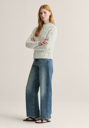 Ung kvinde med rødt hår iført en hvid kabelstrikket sweater, blå viddebukser og sorte sandaler, stående med armene krydsede.