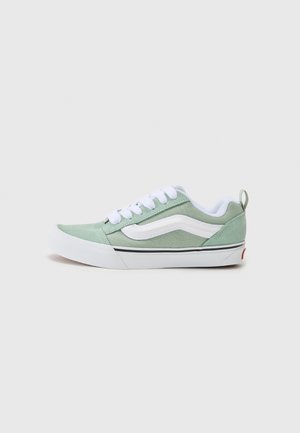 Green canvas sneaker with white accents, round toe, and white laces. Features black trim and a tab at the heel. Flat rubber sole.