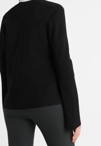 Black structured blazer with a modern fit, featuring long sleeves and a smooth texture, paired with dark green fitted pants.