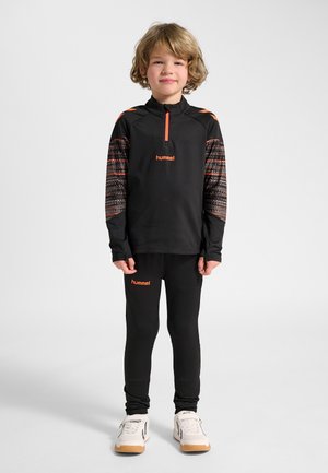 Child standing wearing black sportswear with orange accents and white sneakers against plain white background.