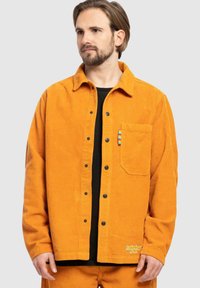 Corduroy jacket in bright orange with black buttons, a chest pocket, and a multicolored accent. Pairing with black shirt underneath.