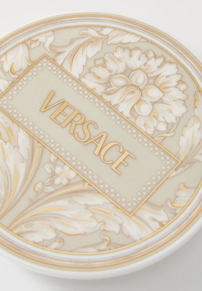 Bílý keramický talíř zdobený složitými květinovými vzory v béžové a zlaté barvě, s centrálním obdélníkovým panelem nesoucím nápis "VERSACE" tučným písmem.