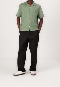 Camisa verde de manga corta con un bolsillo y cierre de botón, combinada con pantalones negros de corte holgado y zapatillas blancas con acentos beige.