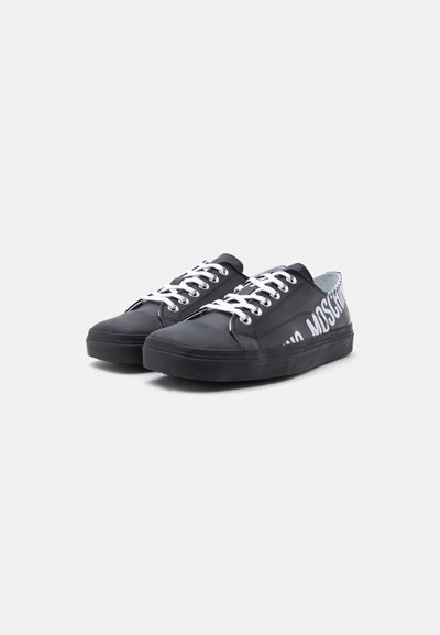 Ζευγάρι μαύρα sneakers χαμηλού τύπου με άσπρα κορδόνια και έντονα λευκά γράμματα στο πλάι, πάνω σε απλό ανοιχτό φόντο.