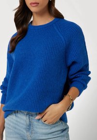 Niebieski sweter dzianinowy z fakturowanym wzorem rib, okrągłym dekoltem i luźnym krojem. Połączony z jasnoniebieskimi dżinsami i złotą bransoletką.