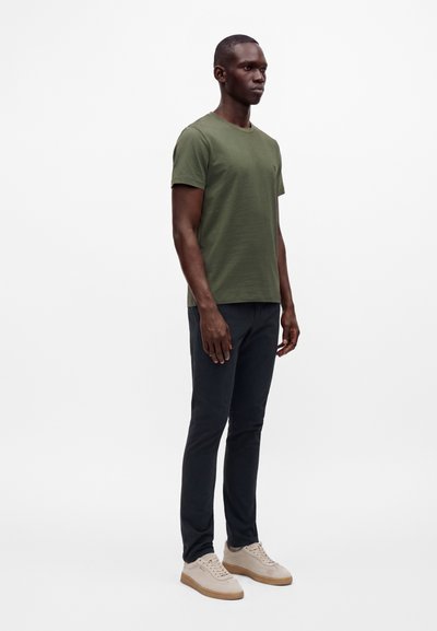 Homme debout portant une chemise verte à manches courtes, un pantalon sombre et des baskets beiges sur un fond blanc uni.