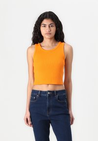 Skraćena narančasta rebrasta majica bez rukava sa širokim naramenicama, u kombinaciji sa tamnim trapericama visokog struka koje imaju klasične džepove i ukrase od šivanja.