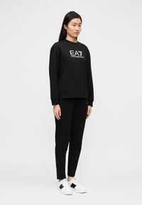 Σετ μαύρη φόρμα με μπλούζα και παντελόνι, κατασκευασμένο από απαλό βαμβάκι, με λογότυπο στο μπροστινό μέρος. Συνδυάζεται με λευκά αθλητικά παπούτσια που έχουν μαύρες λεπτομέρειες.