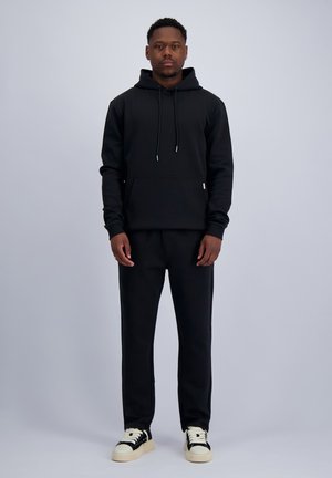 Mann steht frontal, trägt einen schwarzen Hoodie, schwarze Hose und schwarz-weiße Sneaker vor einfarbig hellem Hintergrund.