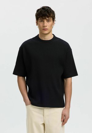 Ung mann med krøllete hår som har på seg en svart ribbestrikket kortermet skjorte og beige bukser, står mot en ensfarget lys bakgrunn.