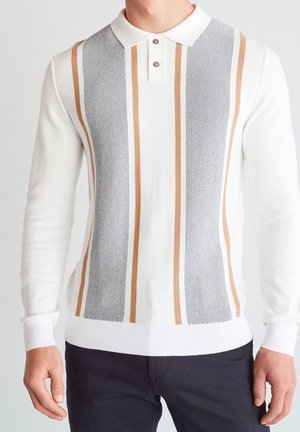 Homme portant un polo blanc à manches longues avec des rayures verticales beige et noires à pois, associé à un pantalon sombre, debout devant un fond uni.