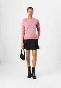 Pink crewneck sweatshirt with ribbed cuffs, paired with a black pleated skirt. Black ankle boots and a small black handbag complete the look.