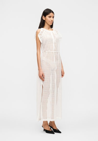 Γυναίκα που φορεί ένα λευκό μακρύ φόρεμα από κροσέ χωρίς μανίκια με πλαϊνή σκίσιμο, σε συνδυασμό με μαύρες μυτερές γόβες, μπροστά σε απλό λευκό φόντο.