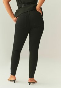 Juodos siaurėjančios džinsinės kelnės, pagamintos iš džinso audinio, su vidutinio aukščio liemeniu ir penkių kišenių dizainu. Modelis dėvi juodus atvirų pirštų aukštakulnius.