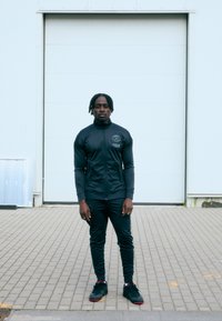 Sportlane tumedates hallides spordirõivastes, kõrgema kraega ja logoga, seismas ühtlase valge seina ees, jalas mustad spordijalatsid.