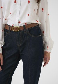 Jeans hlače iz jeansa z oranžnim šivom, kombinirane z belo bluzo z vezenino rdečih vrtnic. Rjav usnjen pas z bronasto zaponko.