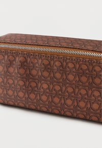 Trousse de maquillage marron avec fermeture éclair, présentant un motif géométrique en relief, une forme rectangulaire élégante et des détails en métal.