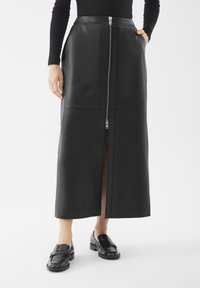Vrouw draagt een zwarte leren midi-rok met een ritssluiting aan de voorkant en zakken, gecombineerd met zwarte instappers en een zwart T-shirt met lange mouwen.