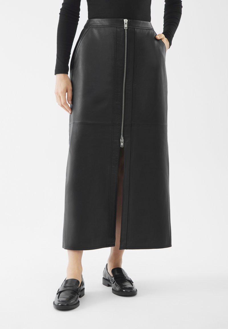 Vrouw draagt een zwarte leren midi-rok met een ritssluiting aan de voorkant en zakken, gecombineerd met zwarte instappers en een zwart T-shirt met lange mouwen.
